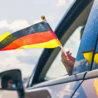 Main sortant d’une voiture tenant un petit drapeau allemand, illustrant une actualité liée à l’Allemagne.