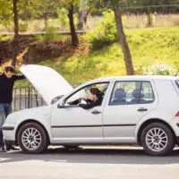 Conducteur devant une voiture immobilisée capot ouvert sur le bord de la route, illustrant un imprévu mécanique et la nécessité de sécuriser la situation.